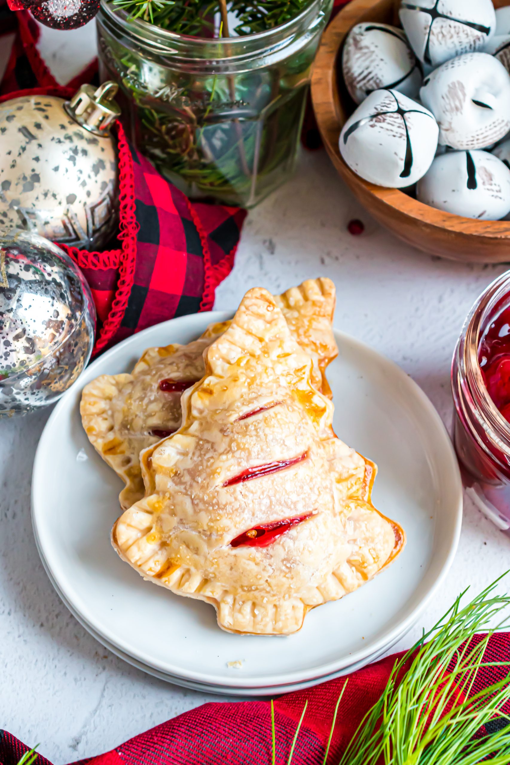 A tree shaped cherry hand pie on a white plate surrounded by christmas ornmaments and greenery.