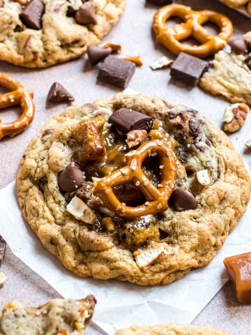 A kitchen sink cookie laying on a piece of parchment paper with caramel, chocolate and pretzel bits scattered around it.
