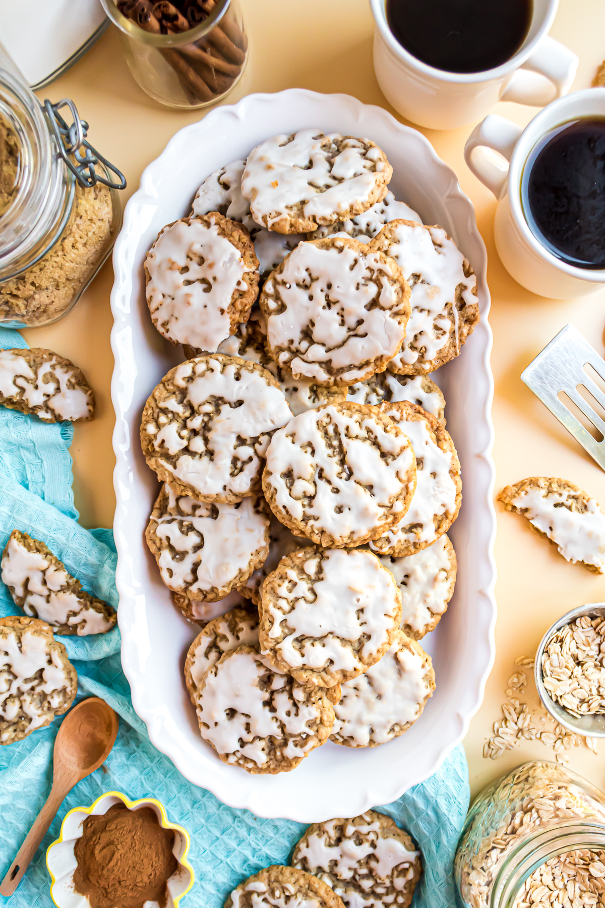 There is an arrangement of iced oatmeal cookies without egg on a white platter sitting on a yellow background with an aqua blue linen tucked under one of the edges. There are broken oatmeal cookies scattered about the surface and an open jar containing brown sugar in the upper left-hand corner. A cup of coffee, a serving spatula, a saucer of cinnamon, and a small dish of oats also frame the serving dish.