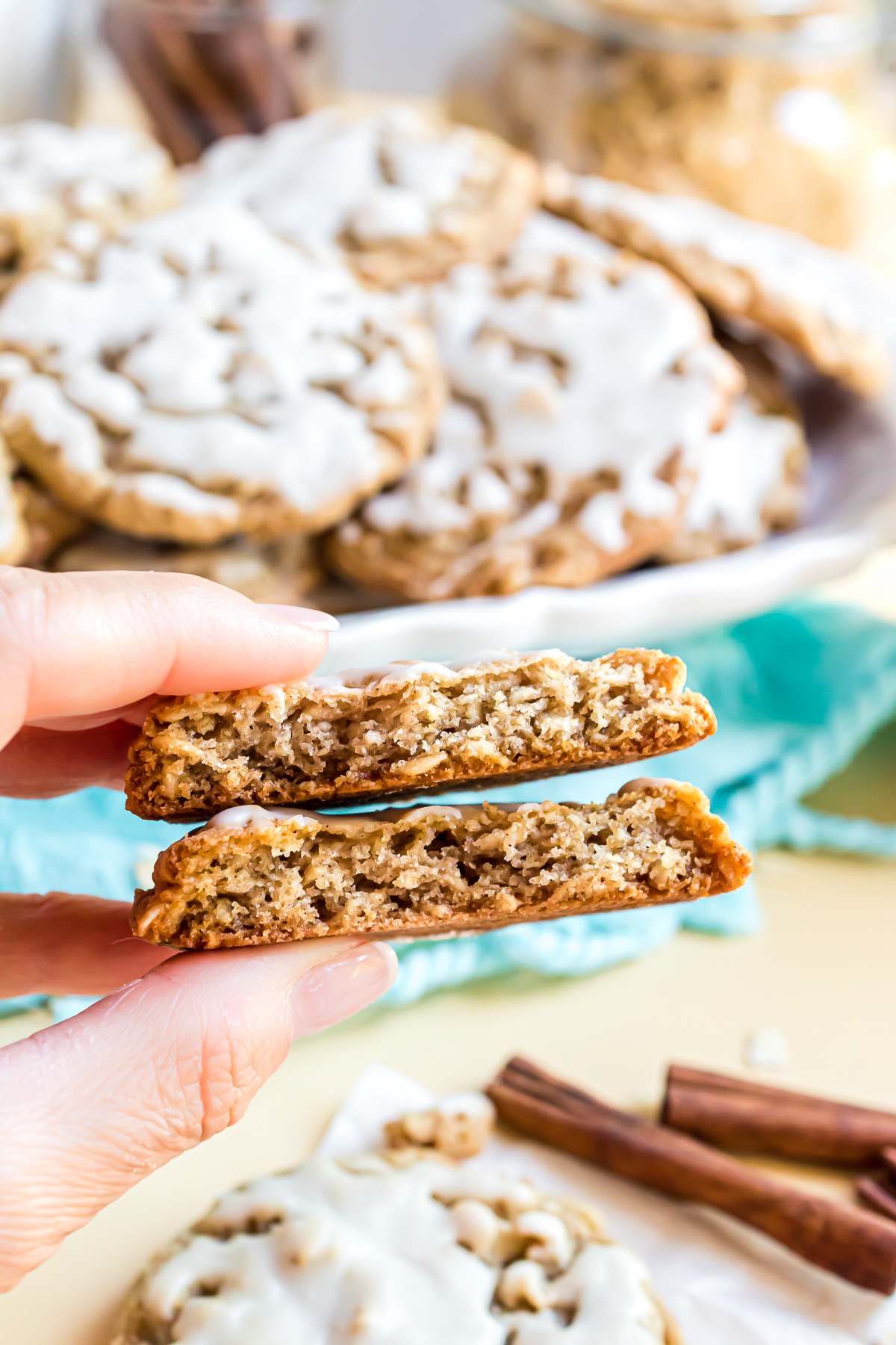 An iced oatmeal cookie broken in half and stacked on top of each other to show the inside texture of the cookie. It is golden brown, pieces of oats can be seen inside the cookie, and a little icing is peeking out from the cookies' tops. A platter of cookies is in the background.