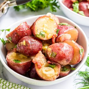 A serving of warm potato salad in a white bowl. The potato salad is coated with the mustard dressing and topped with bacon and green onions.