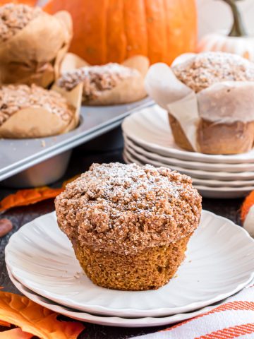 A muffin made with the Panera Bread Pumpkin Muiffin recipe sits on a white plate with a tin full of muffins and a large pumpkin behind it.