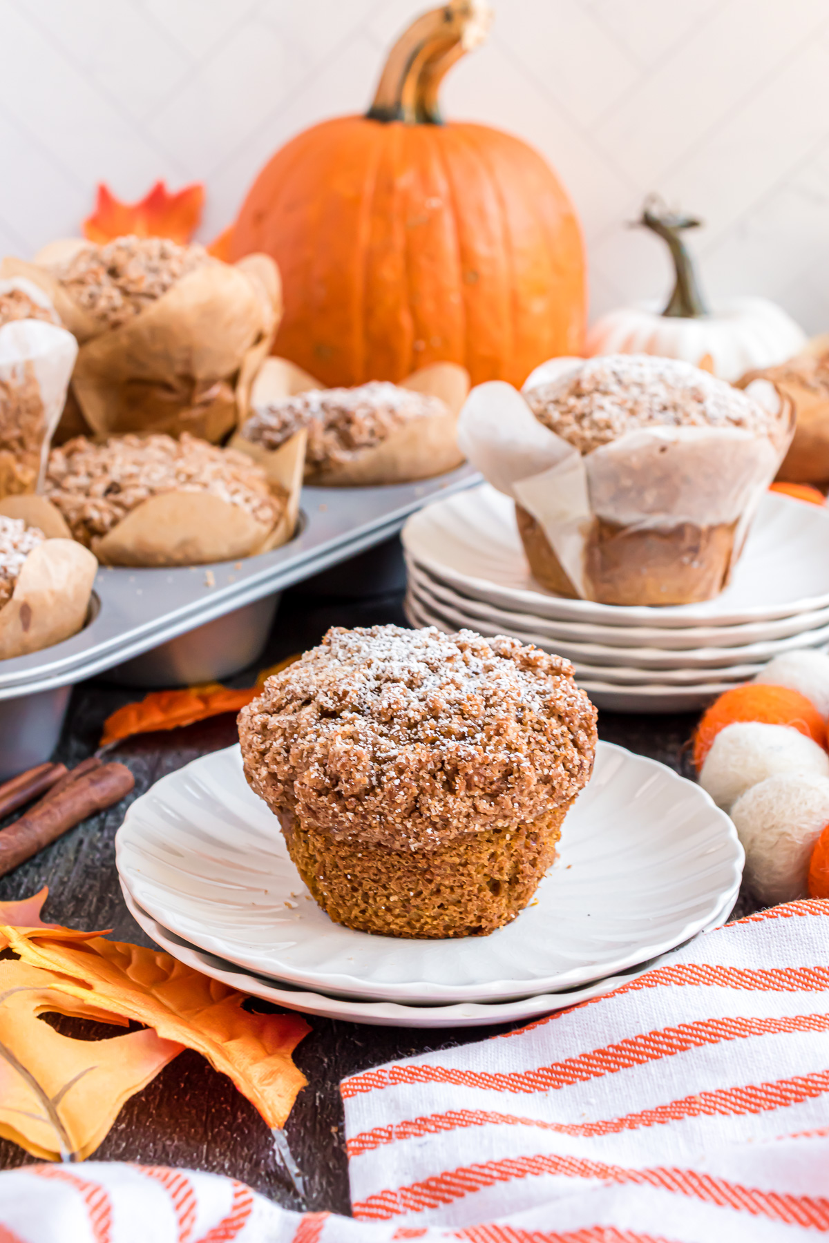 A muffin made with the Panera Bread Pumpkin Muiffin recipe sits on a white plate with a tin full of muffins and a large pumpkin behind it.