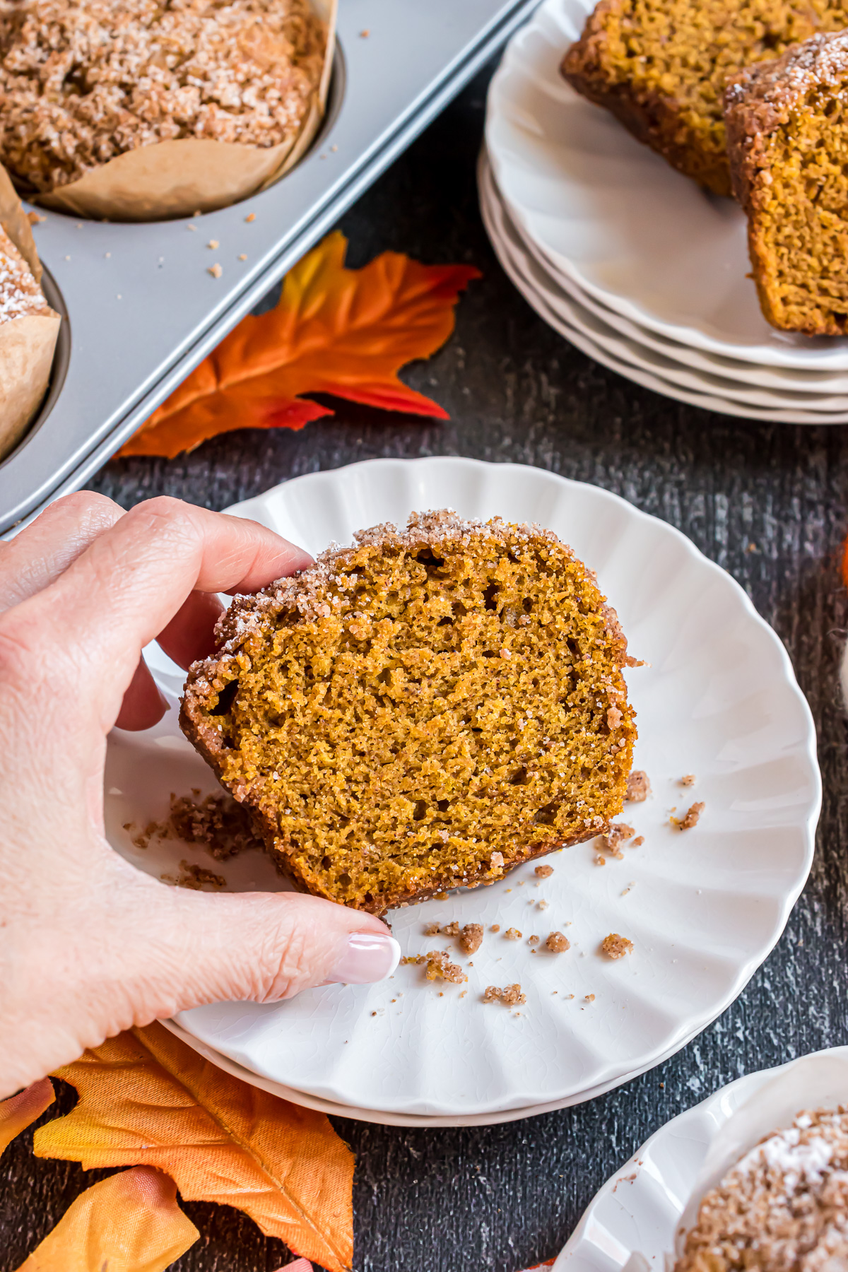 A white scalloped edge plate with half of a pumpkin muffin on it sits on a dark grey wood background. The plate is dotted with streusel crumb topping that has fallen off the muffin. A hand is reaching in from the bottom left hand corner and grabbing the muffin.