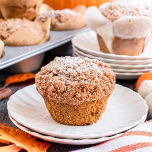 A pumpkin muffin on a plate with the wrapper removed. There is a tin full of muffins and another plated muffin in the background.