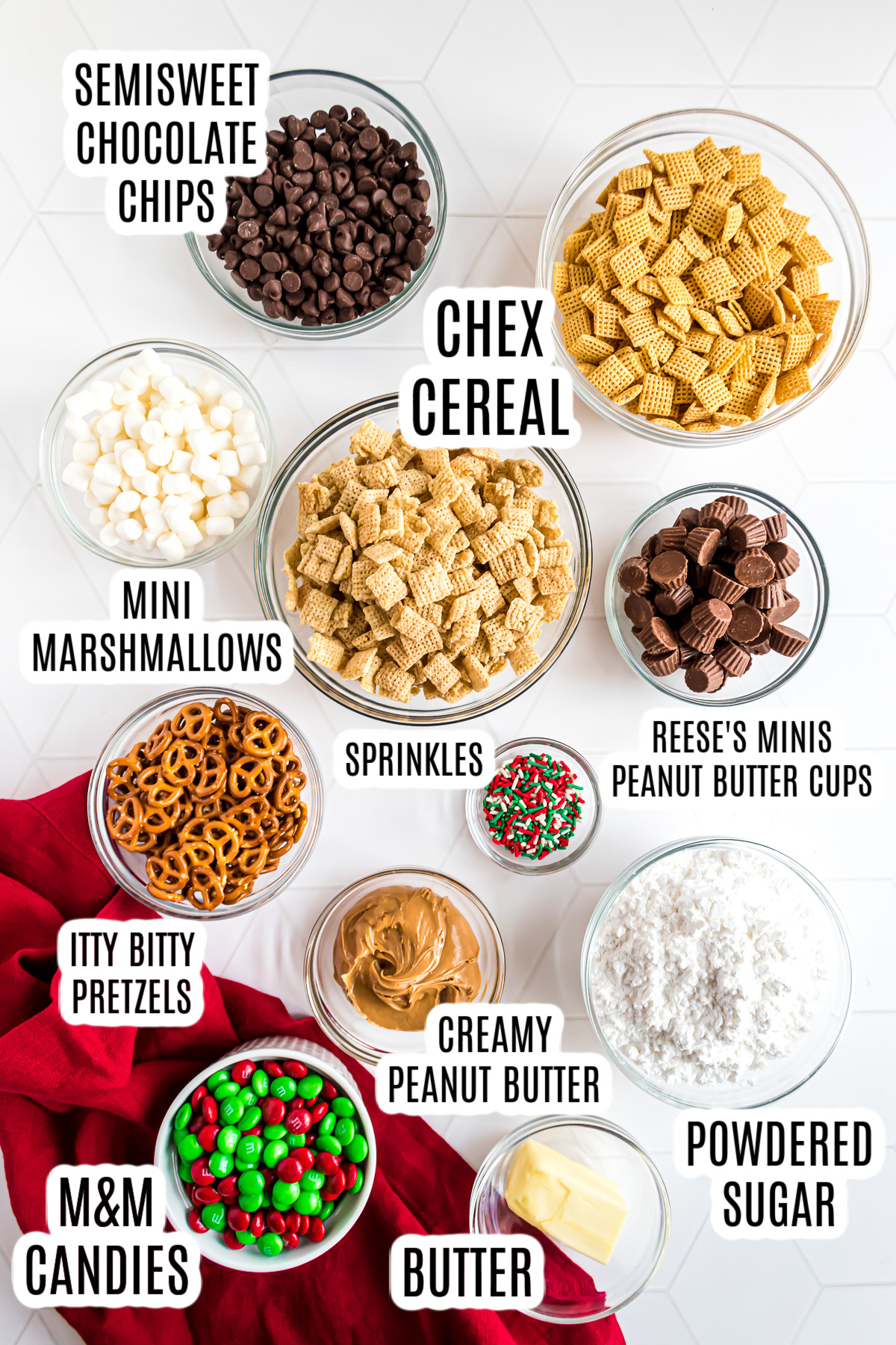 All the ingredients needed to make the Christmas Reindeer Chex Mix Recipe including: Chex cereal, semisweet chocolate chips, mini marshmallows, holiday sprinkles, Reese's Minis peanut butter cups, pretzels, creamy peanut butter, powdered sugar, red and green m&m candies, and butter.