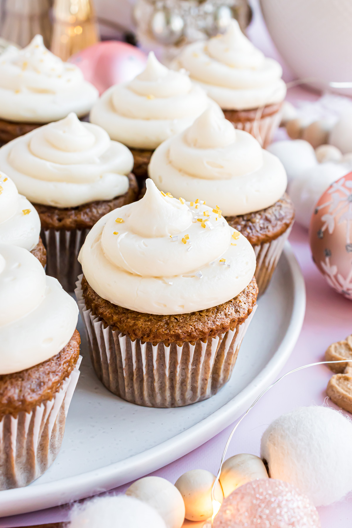 A gingerbread cupcake on a white serving platter topped with a swirl of cream cheese frosting, adorned with gold and silver sprinkles and surrounded by other cupcakes.