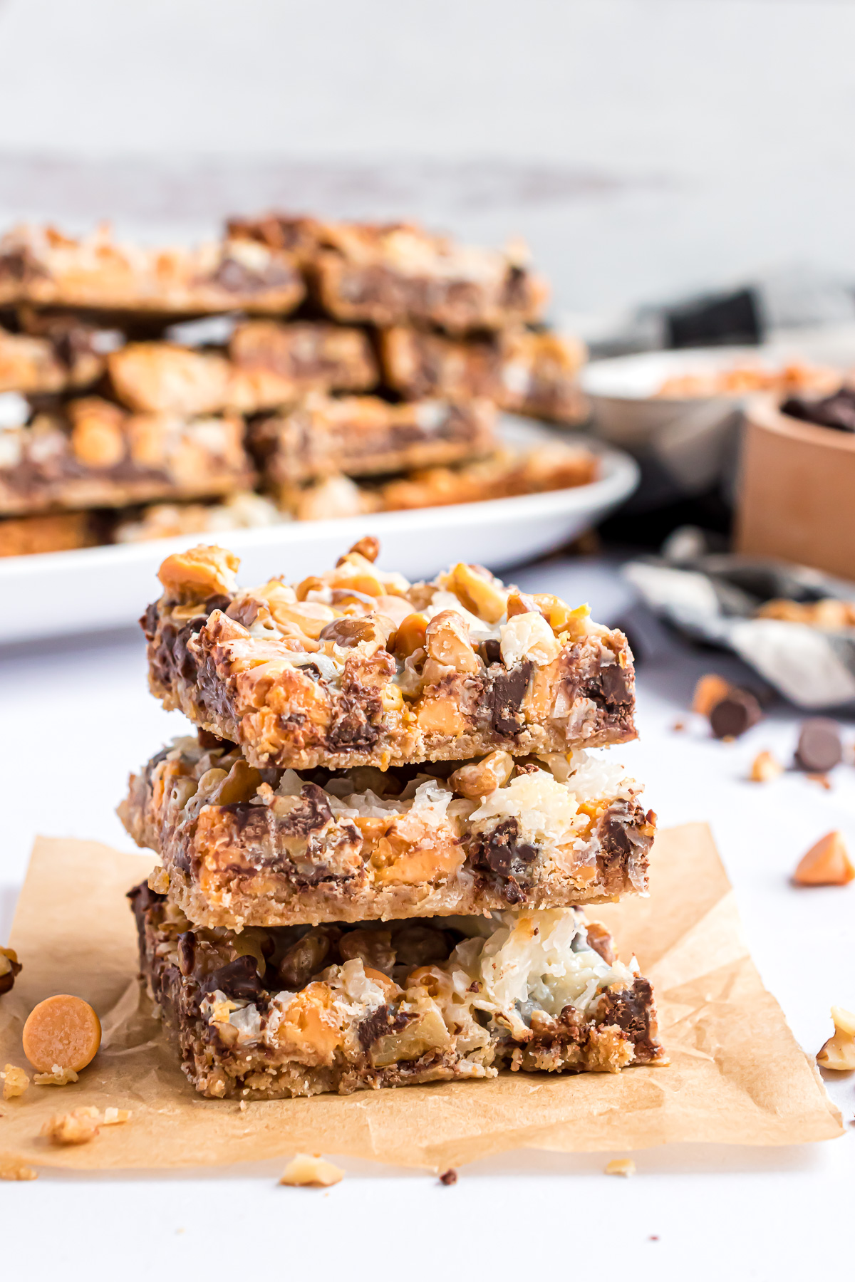 A stack of 3 cookies, sitting on a piece of brown parchment paper with butterscotch and semi-sweet chocolate chips scattered around the stack. A platter of dolly bars is sitting in the background.
