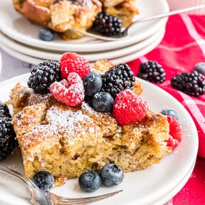 A piece of french toast casserole topped with powdered sugar and berries and plated with a fork resting on the plate.