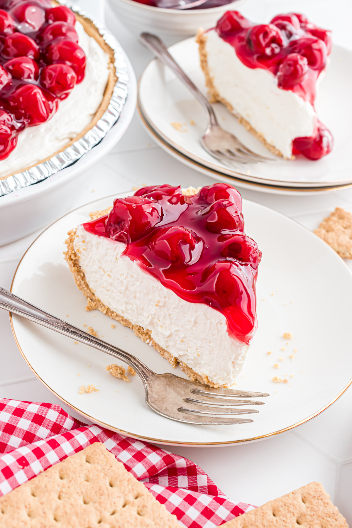 A piece of cherry cheesecake pie on a plate with a fork. There is another plated piece of dessert in the background. 