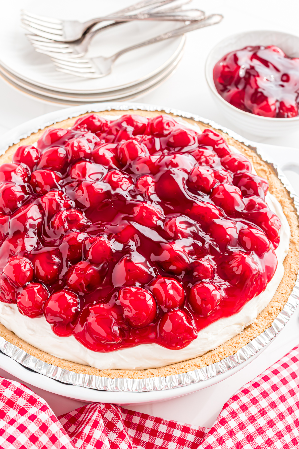 A pie plate with a No Bake Cherry Pie. There is a stack of small dessert plates in the background and a dish of cherry pie filling.