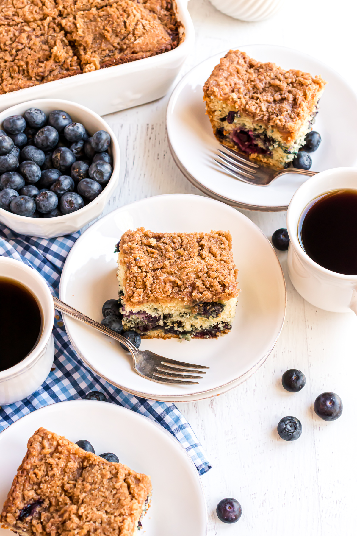 There are 3 pieces of blueberry dessert cake served on individual plates. They are surrounded by fresh blueberries on each plate and cups of coffee.