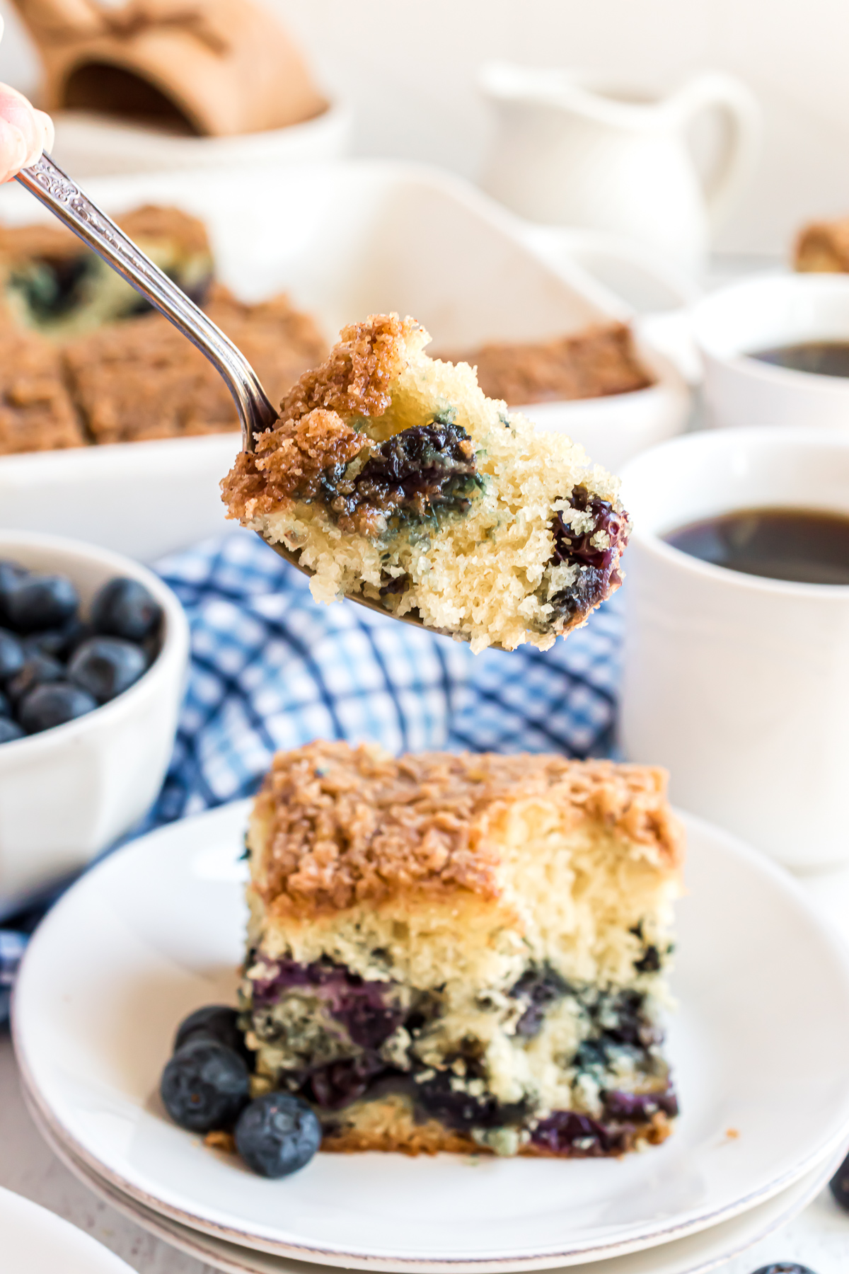 A bite of the blueberry coffee cake on a fork with the cake piece shown below the bite and two cups of coffee in the background.
