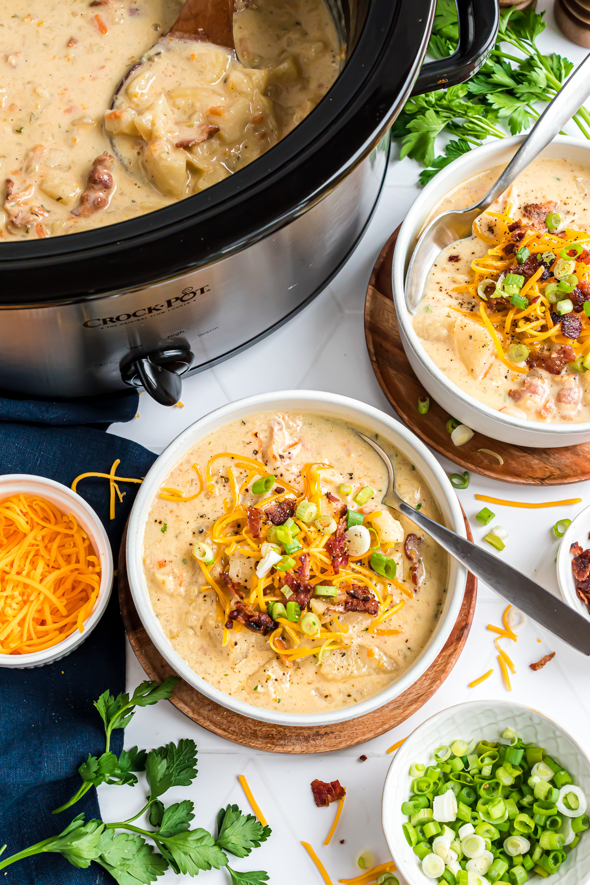 A bowl of creamy potato soup topped with sliced green onions, crumbled bacon and cheddar cheese. A crockpot full of soup is in the upper left-hand corner and another bowl of soup fills the upper right-hand corner.