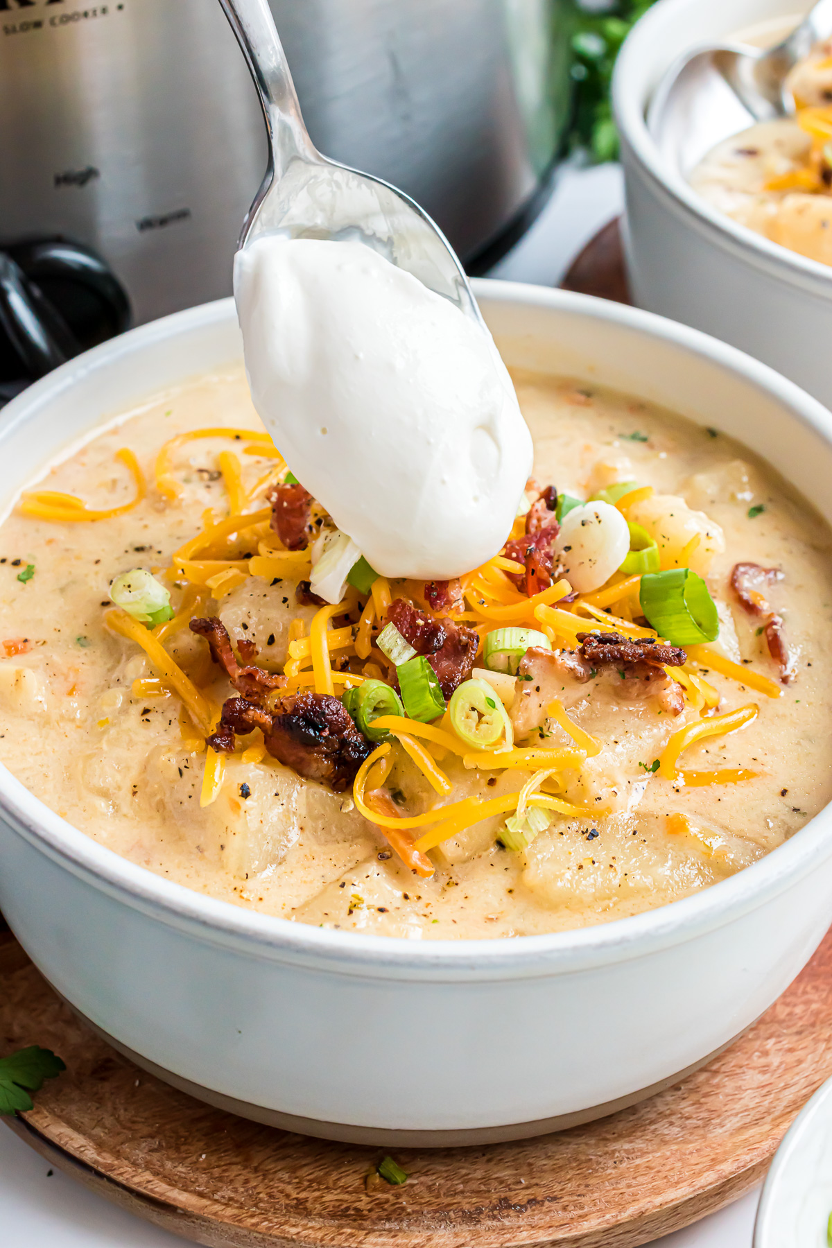 A bowl filled with baked potato soup topped with a garnish of bacon, green onions, and cheese and a dollop of sour cream being spooned onto the top of the soup.