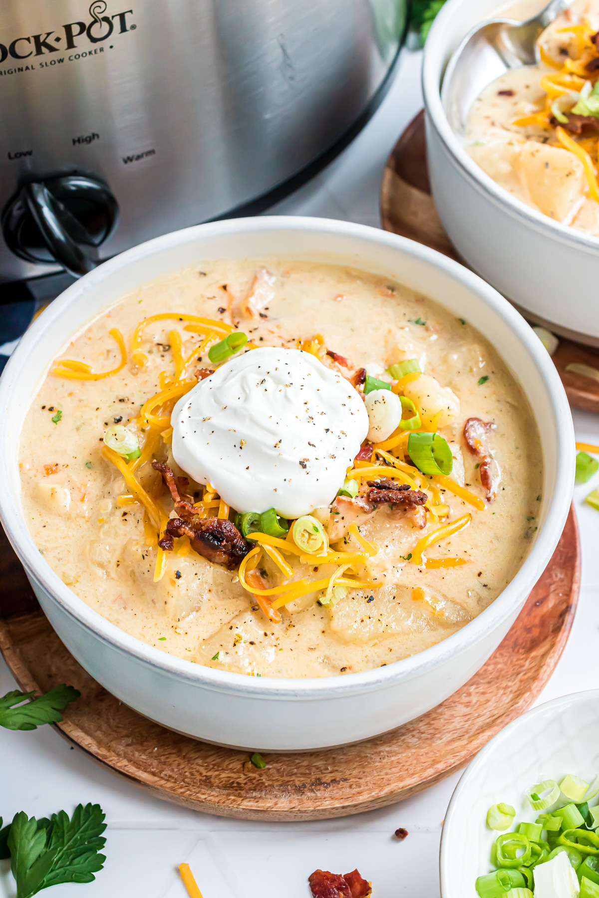 A bowl of loaded baked potato soup sitting on a wooden saucer topped with sour cream and ground black pepper.