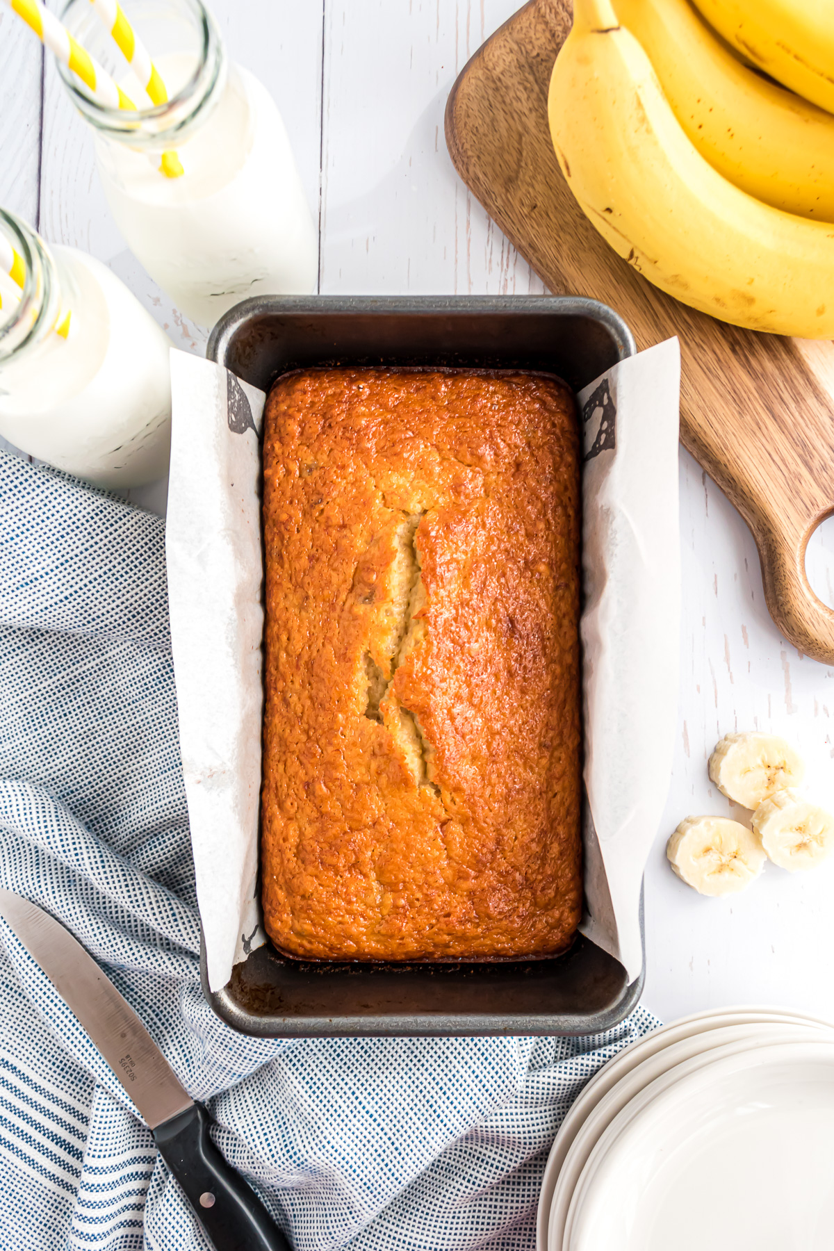 An overhead view of a loaf of banana bread in the pan on a white plank background with two glasses of milk and a bunch of bananas on a wooden cutting board in the upper right-hand corner.