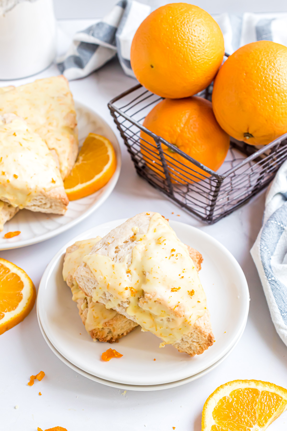 Two copycat panera bread glazed orange scones on a plate surrounded by orange slices and a basket of oranges and a serving plate full of scones in the background.