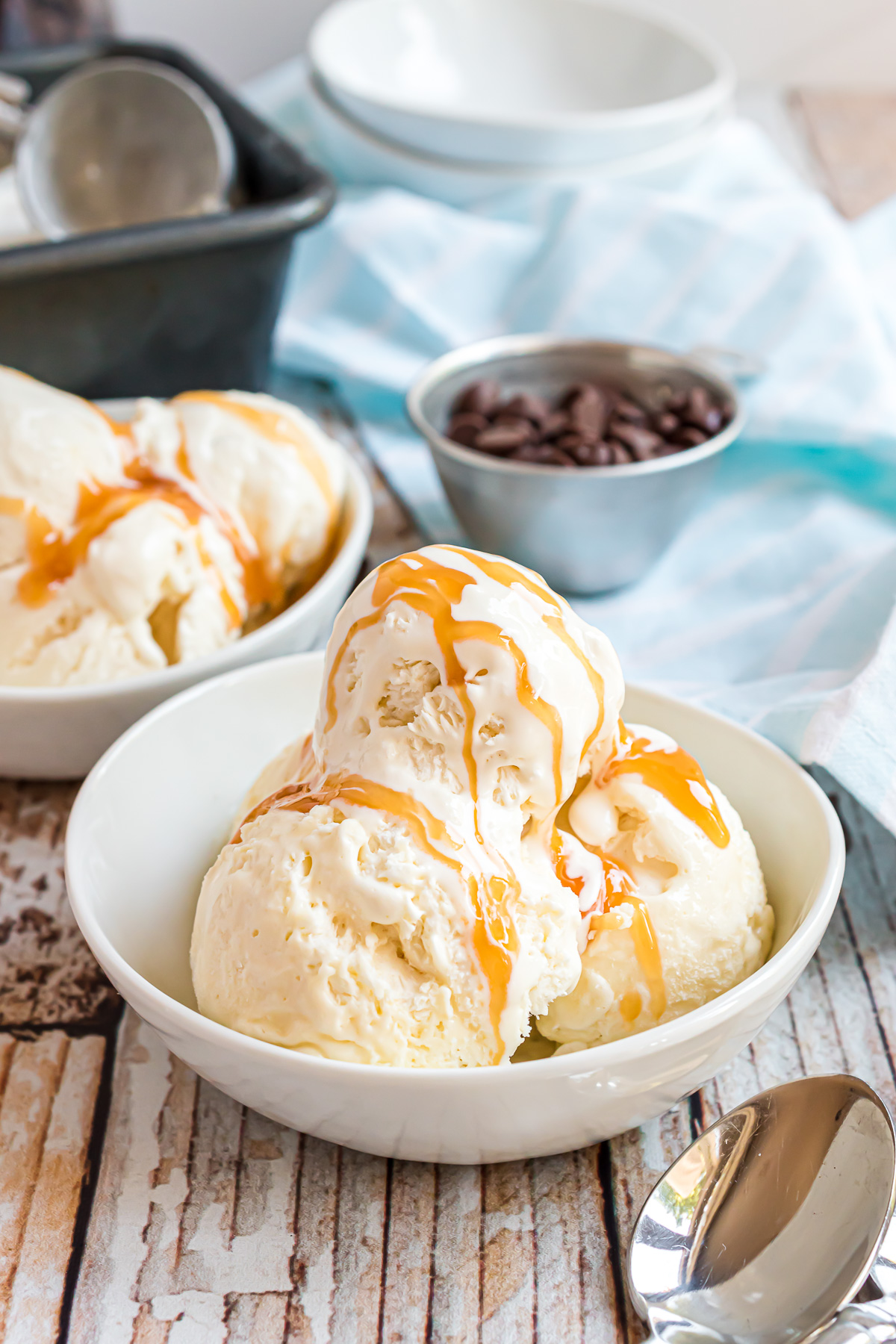 A white ceramic bowl with three large scoops of vanilla ice cream in it. The ice cream is drizzled with caramel sauce and the ice cream is melting and creamy.