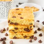 A stack of three Chocolate Chip Cookie Bars on a white plate with chocolate chips scattered around them.