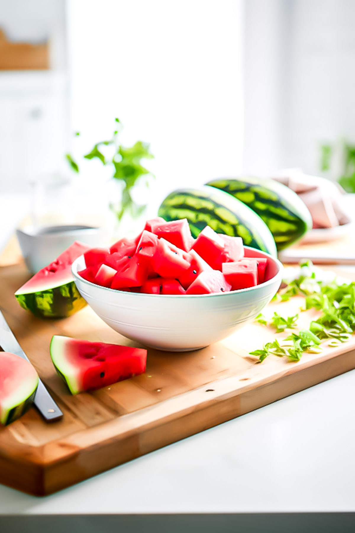A bowl of watermelon that has been cut into large chunks, sitting on a wooden cutting board.