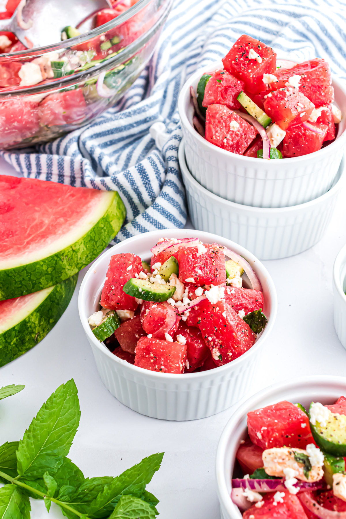 Three small bowls of watermelon salad topped with crumbled goat cheese, slices of red onion and cucumber wedges.