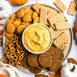 A round serving platter with a small dish of pumpkin fluff in the center surrounded by vanilla wafers, graham crackers, pretzels, and ginger snaps.