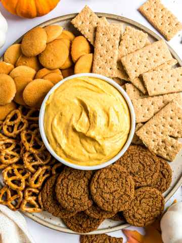 A round serving platter with a small dish of pumpkin fluff in the center surrounded by vanilla wafers, graham crackers, pretzels, and ginger snaps.