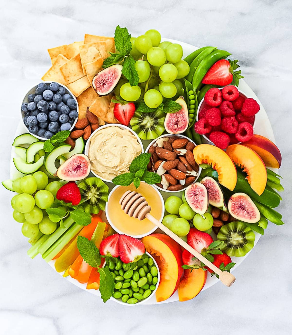 A vegertarian charcuterie board featuring vibrant fresh produce including raspberries, grapes, kiwis, blueberries, cucumbers, peaches, celery and much more.