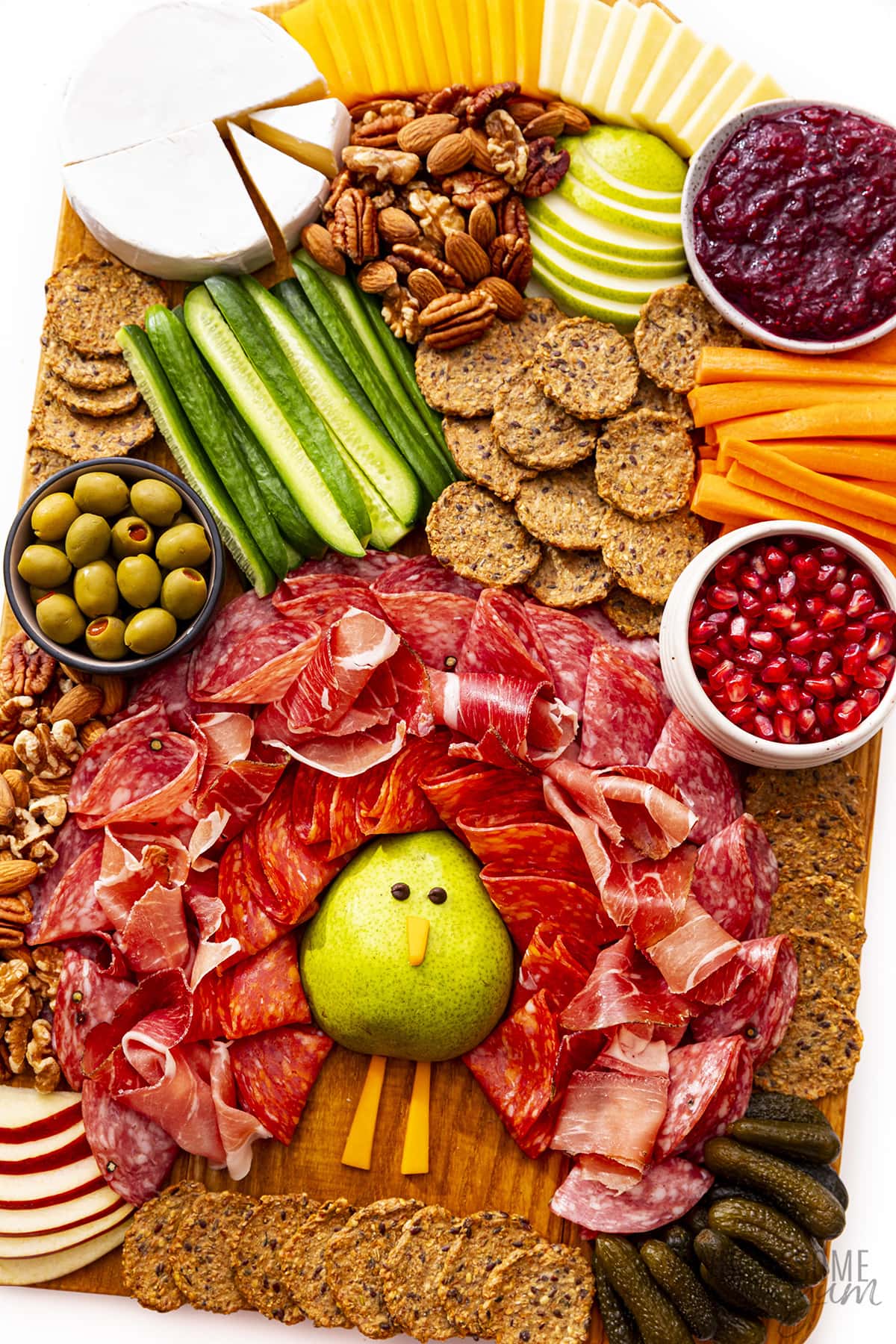 A meat and cheese board with a turkey made from half a pear surrounded by folded meat for the tail feathers. The board is accented by crackers and fresh veggies.