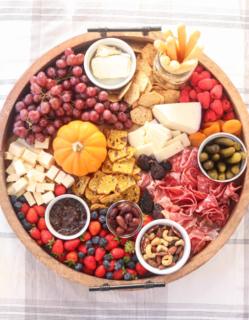 A round snack platter with pickles, nuts, fruit, cheese and meats.