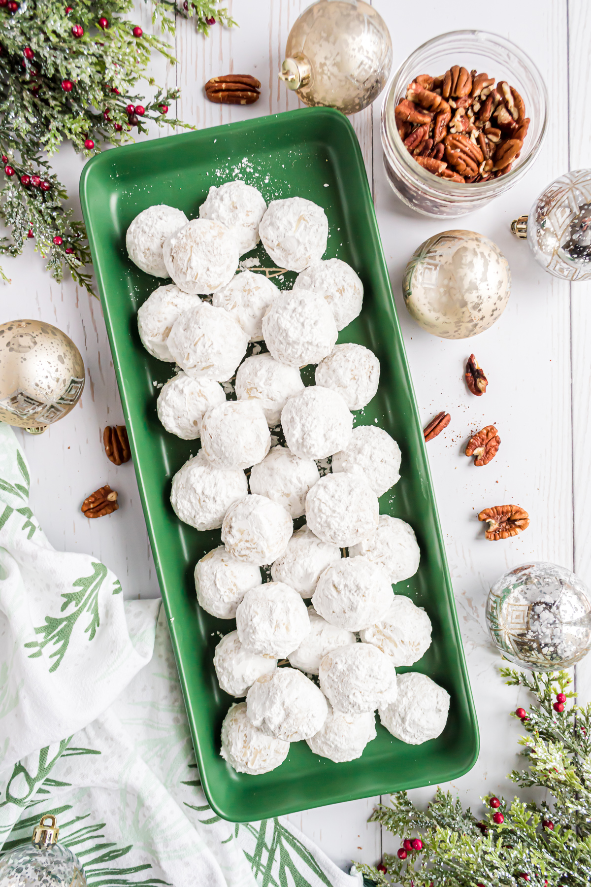 A green rectangle tray of snowball cookies surrounded by holiday ornaments, greenery and pecans.