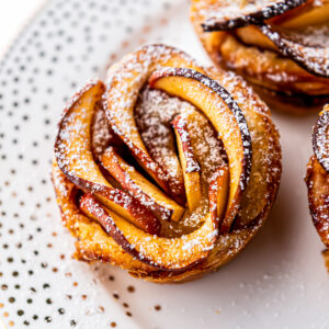 An Apple Rose with Puff Pastry sprinkled with powdered sugar on a white plate.