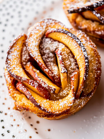 An Apple Rose with Puff Pastry sprinkled with powdered sugar on a white plate.