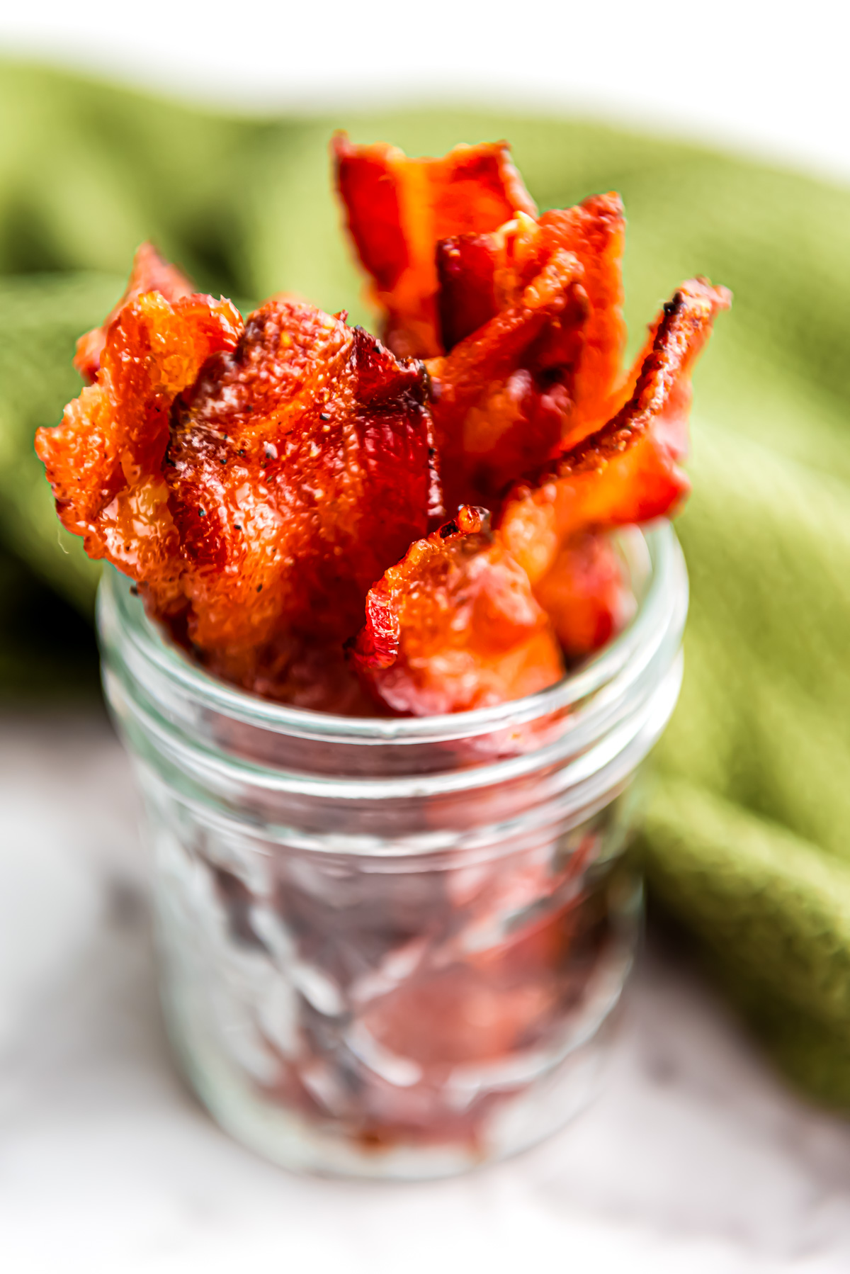 A glass mason jar with strips of candied maple bacon standing upright to show the crispy texture and the caramelized maple coating,