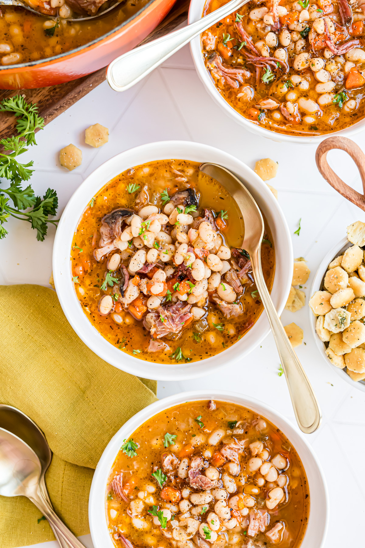 Three bowls filled with delicious navy bean and ham soup on a white tile countertop surrounded by fresh parsley and a scattering of seasoned soup crackers.