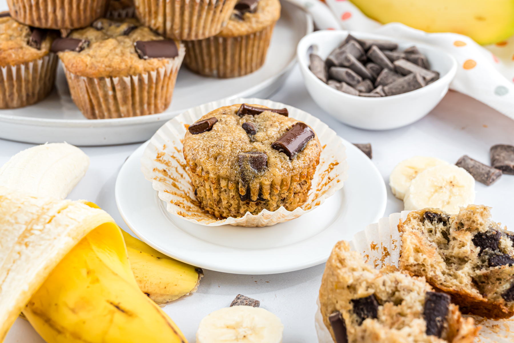 A Chocolate Chunk Banana Muffin on a plate with a large serving platter full of muffins behind it on the countertop.