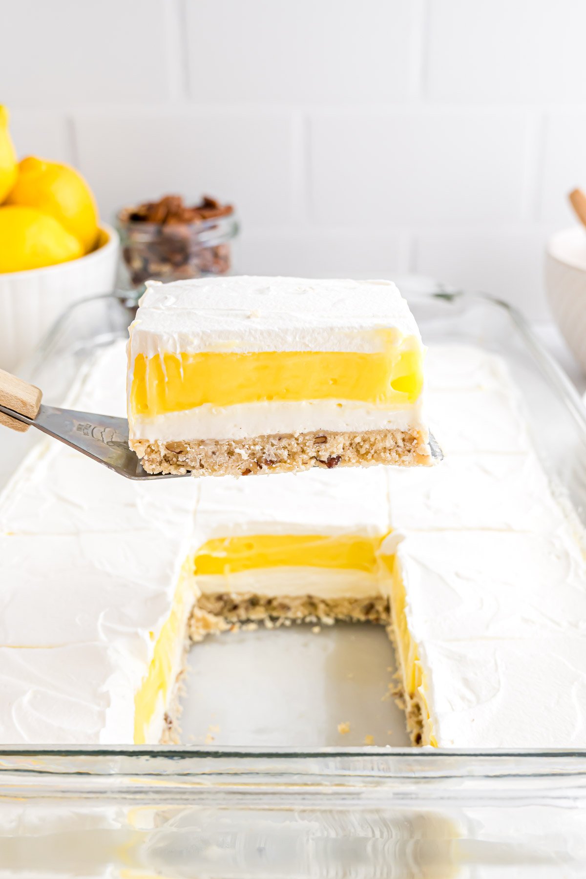A piece of Lemon Lush that has been cut and it being lifted out of a glass baking dish on a spatula showing the distinct layers of the dessert - crust, cream cheese layer, lemon pudding, and Cool Whip topping.