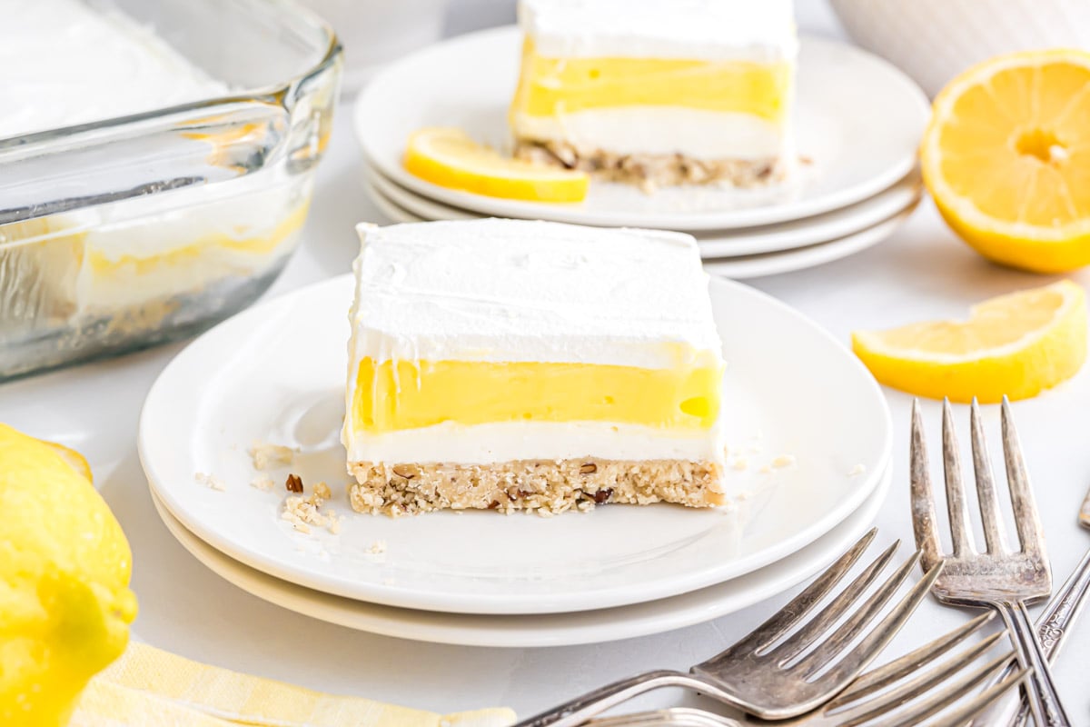 A piece of Lemon Lush dessert on a small serving plate. In the background is another piece on a plate and the 9" x 13" dish with the rest of the layered dessert in it.