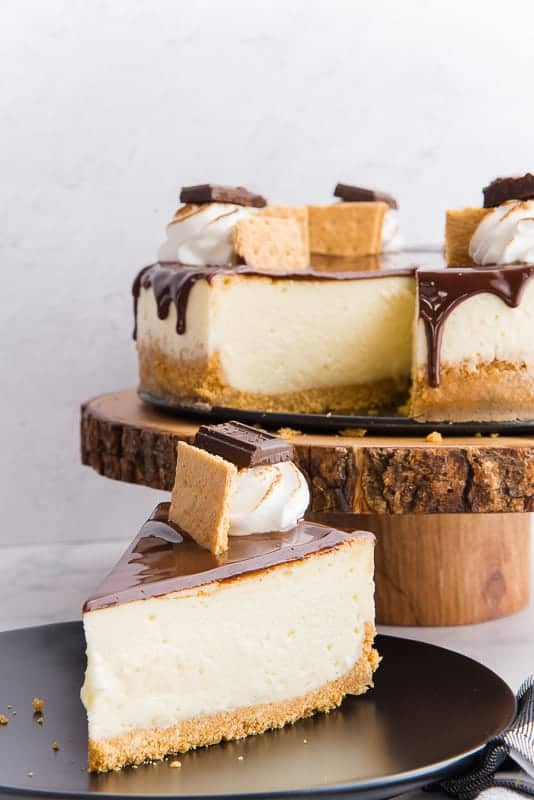 A S'mores cheesecake on a cake stand with a piece of cheesecake on a small serving plate in the foreground.