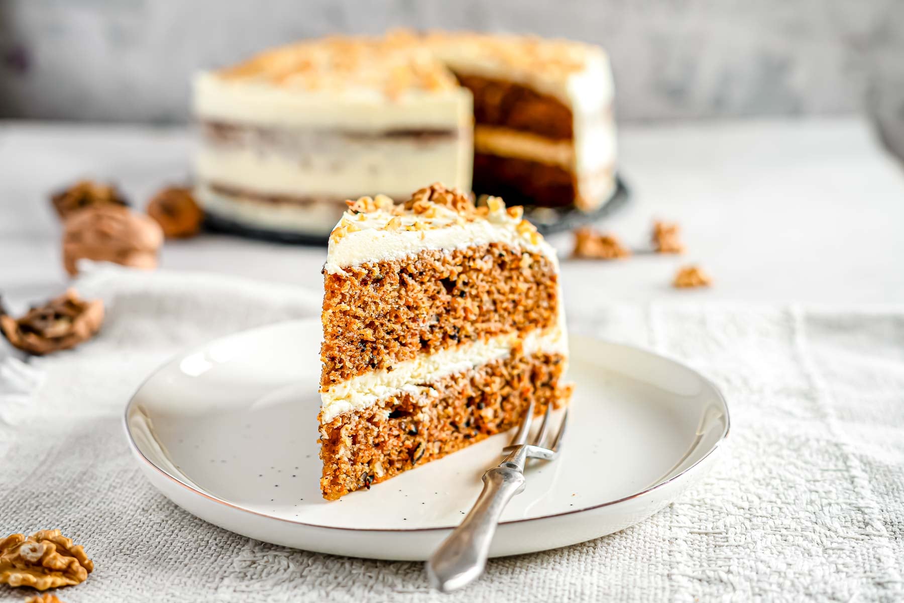 A piece of carrot cake on a plate with a fork. There is a full 2-layer carrot cake in the background.