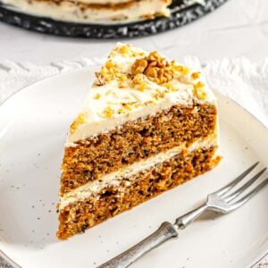 A piece of layered carrot cake topped with cream cheese frosting and nuts on a small serving plate.
