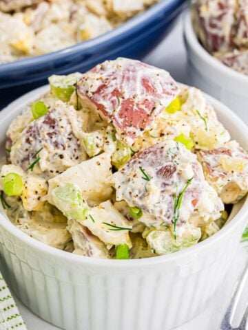A serving of red potato salad with a creamy dressing, bits of celery, dill pickle, onion, and fresh dill in a ramekin.