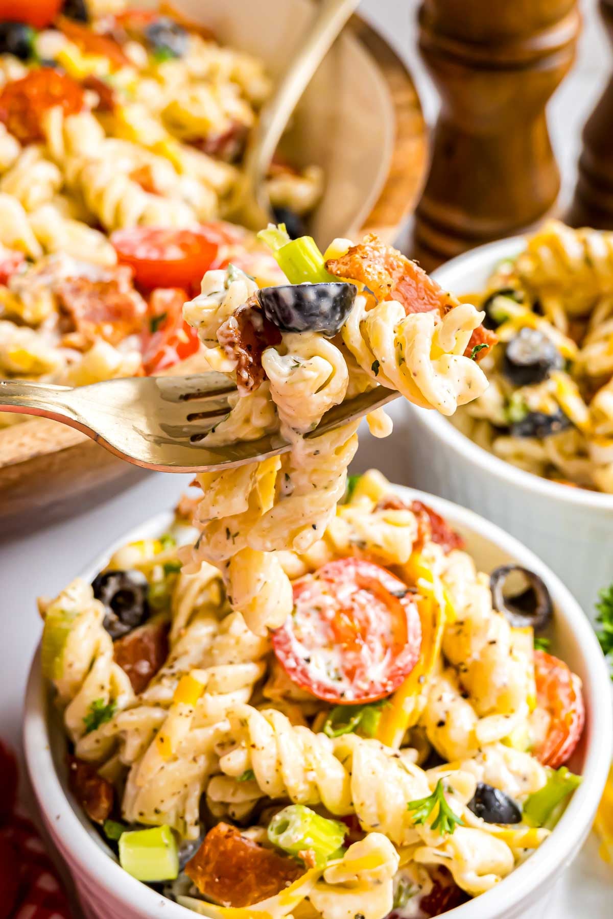 A close-up, vibrant shot of a forkful of creamy Bacon Ranch Pasta Salad, highlighting a perfect bite of rotini coated in homemade ranch dressing with visible pieces of crispy bacon, sliced black olives, and fresh green onions.