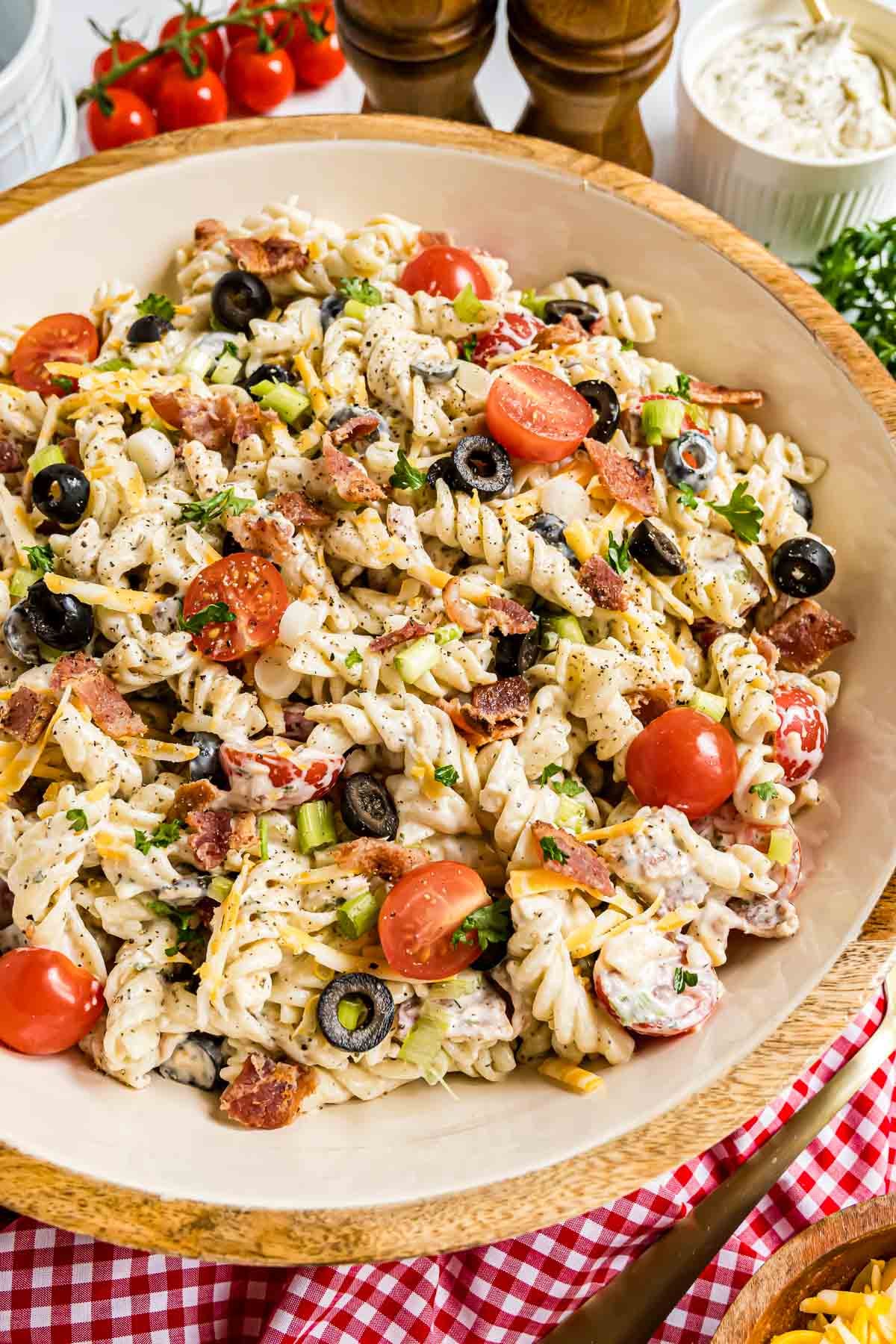 A vibrant, overhead shot of a large serving bowl filled with creamy Bacon Ranch Pasta Salad, showcasing perfectly cooked rotini tossed with crispy crumbled bacon, bright cherry tomatoes, sliced black olives, shredded Colby Jack cheese, and fresh green onions, all generously coated in a smooth, homemade ranch dressing.